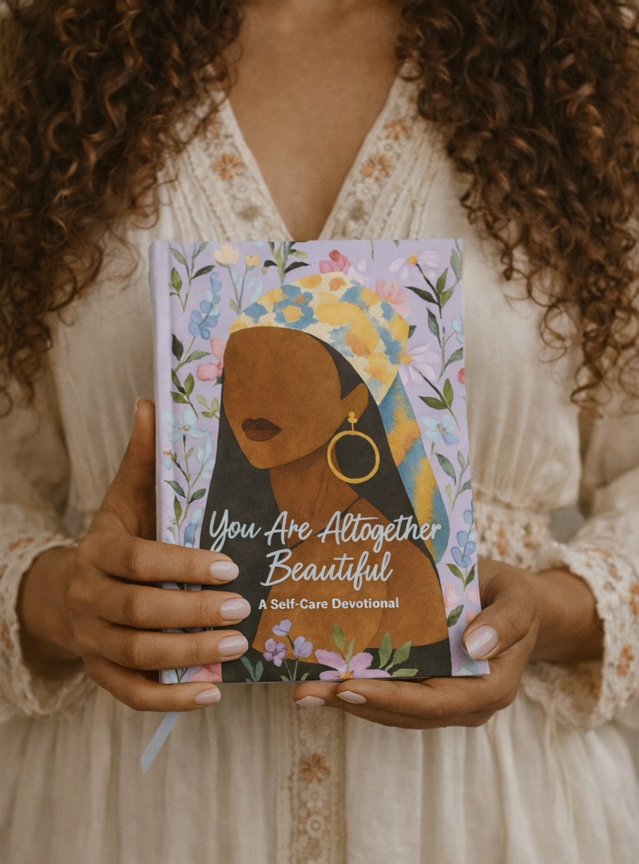 A person in a cream lace dress holds a decorative book titled 'You Are All Together Beautiful: A Self-Care Devotional'. The book cover features a colorful floral illustration of a woman's face and profile, with the title in white script.